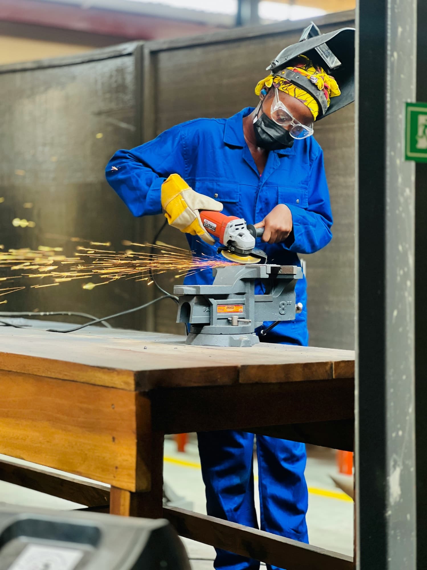 A young woman in a blue work suit and yellow headwrap grinding metal in a workshop in Accra, sparks flying